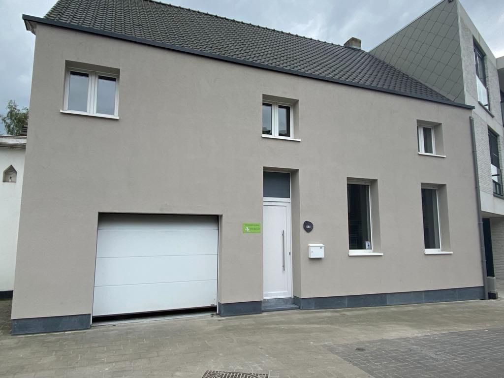 Woninggevel met lichtgrijze crepi, witte garagedeur, witte voordeur en meerdere ramen onder een donker getiled zadeldak.