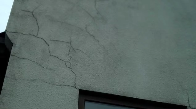 Close-up van een lichtgrijze crepi-gevel met scheuren in het pleisterwerk boven een raamkozijn.